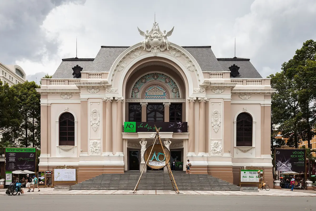 The front of the Saigon Opera House in Ho Chi Minh City, a cream and pink French-colonial building with a wide arched entrance, steps and À Ố Show signs at the door