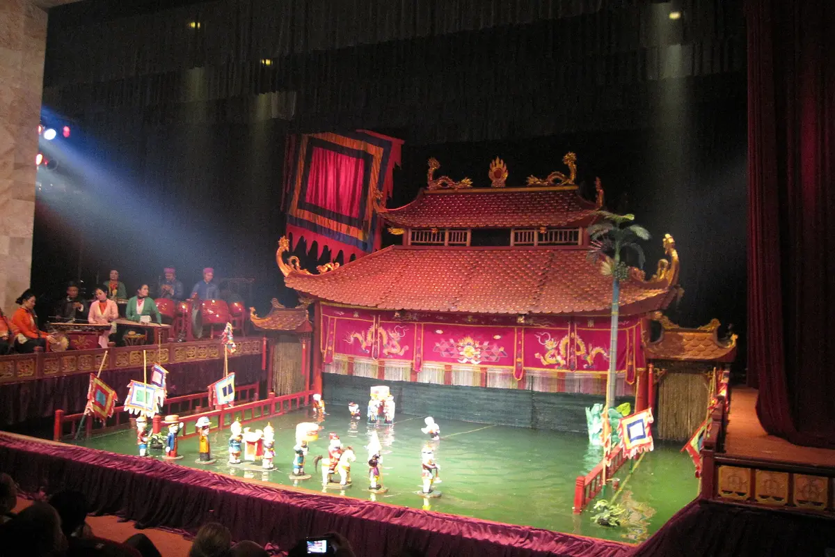 A lit water puppet stage with a red-roofed pavilion at the back, colourful wooden puppets standing on green water and a row of musicians seated on the left